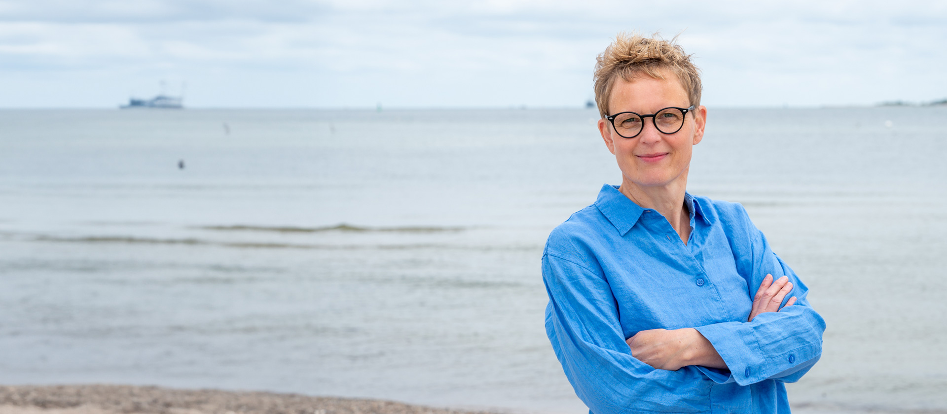 Marlene Rostig mit schwarzer Brille und frechem Kurzhaarschnitt direkt am Ostsee-Strand in Kiel