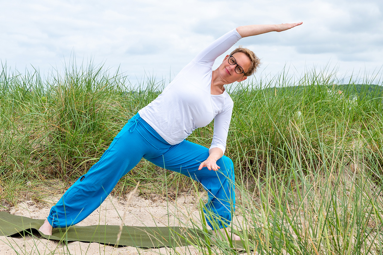 Marlene Rostig auf grüner Yogamatte praktiziert die Yogaübung „gestreckter seitlicher Winkel” in der Dünenlandschaft in Kiel